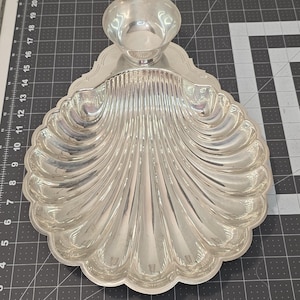 May include: A silver-colored serving dish with a seashell design. The dish has a large, scalloped tray and a small, round bowl attached at the top. The tray has a ribbed texture and a decorative edge. The dish is likely made of metal.