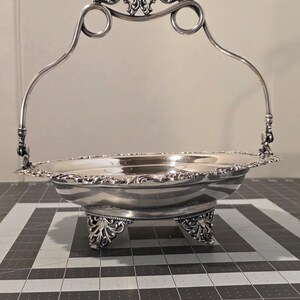 May include: Silver-toned metal serving basket with a handle and ornate detailing. The basket has a round, shallow bowl supported by three decorative feet. The handle features scrollwork and ring accents. A classic piece for serving or display.