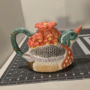 May include: A ceramic teapot shaped like a duck, featuring a green handle with gold accents. The duck's head is green and orange, with a red flower on top. The body is white, gray, and brown.
