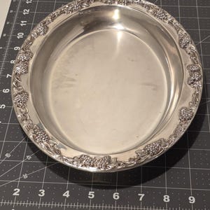 May include: A silver-colored, round serving dish with a decorative border of grape clusters and vines. The dish has a smooth, reflective surface and a slightly raised edge. The dish is likely made of metal.