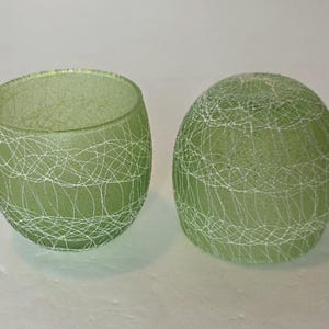 May include: Two green glass candle holders. One is open, the other is a sphere. Both have a white, web-like design. The candle holders are a soft green color and sit on a white surface.