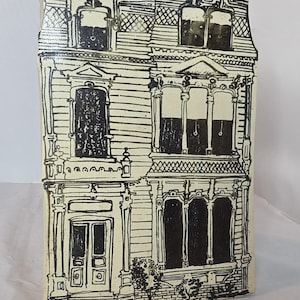 May include: A cream-colored, rectangular ceramic house sculpture with black ink detailing. The artwork depicts a two-story building with windows, doors, and architectural details. The roof has a decorative element at the top.