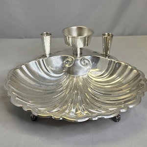 May include: A silver-colored metal serving dish shaped like a large scallop shell. The dish features a central raised bowl and two smaller cylindrical holders on the back edge. It is supported by three small feet and has a polished, reflective finish.