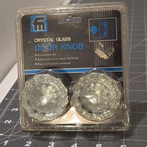 May include: A package containing two crystal glass door knobs. The packaging features the text "CRYSTAL GLASS DOOR KNOB" and states the set is complete, adjustable, and includes instructions. The knobs are clear and faceted.