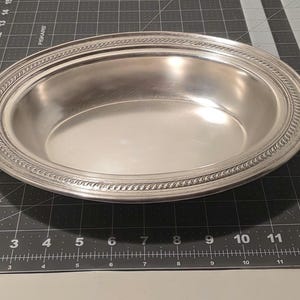 May include: A silver oval serving dish with a decorative rope-like border. The dish is empty and reflects the light, showing a smooth, polished surface. The dish is approximately 12 inches long.
