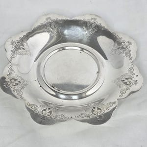 May include: A silver-toned metal dish with a scalloped edge and a circular center. The rim features embossed scrollwork patterns in each of the six lobes. The surface has a reflective, polished finish with a smooth, flat base in the middle.