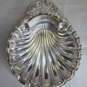 May include: A silver-colored, shell-shaped serving dish. The dish has a scalloped edge and a detailed floral design at the top. The interior has a ribbed texture, and the dish reflects light, creating a shiny appearance.