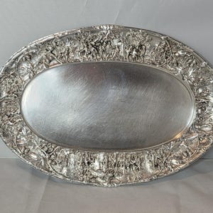 May include: An oval silver-toned metal serving tray featuring a smooth, polished central surface surrounded by a wide, ornate border decorated with detailed relief carvings of figures, trees, and architectural elements in a continuous scene.