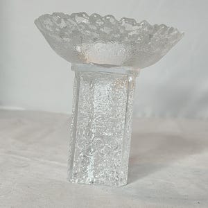 May include: A clear, textured glass candle holder with a ruffled rim bowl and a rectangular base. The base features an embossed floral design. The candle holder is designed to hold a small candle.