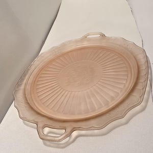 May include: A vintage pink glass serving tray with a scalloped edge and two handles. The tray features a central floral design and radial lines emanating from the center. The tray is approximately 12 inches in diameter.