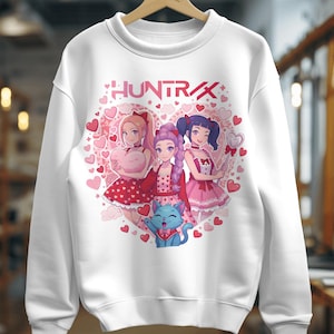 Puede incluir: Sudadera blanca con un diseño en forma de corazón con tres chicas de estilo anime y un gato azul. La palabra "HUNTRAX" se muestra en letras rosas encima de las chicas. El diseño incluye corazones rosas.
