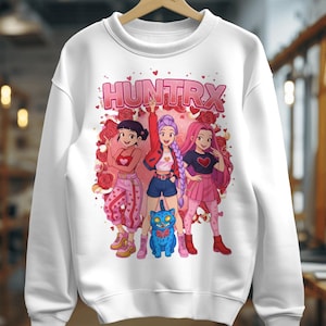 Puede incluir: Sudadera blanca con la palabra "HUNTRX" en rosa sobre un gráfico de tres chicas de estilo anime y un gato azul. Las chicas visten diferentes atuendos, rodeadas de rosas.