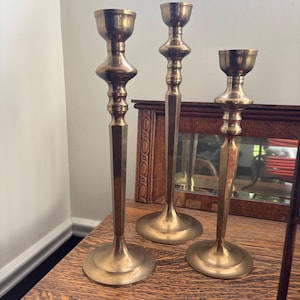 May include: A set of three polished brass candlesticks of varying heights. Each candlestick features a wide circular base, a slender hexagonal stem, and a decorative turned neck leading to a cylindrical cup designed to hold a single taper candle.