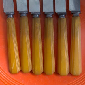 May include: Set of six vintage butter knives. Each knife features a stainless steel blade and a yellow-orange, ribbed handle. The handles have a marbled appearance. The knives are arranged on an orange surface.