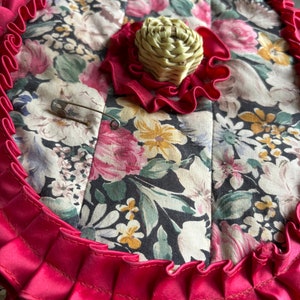May include: A round, quilted fabric box with a floral print in shades of pink, green, and yellow. The box has a red ruffled trim and a woven straw-like knob on the top.
