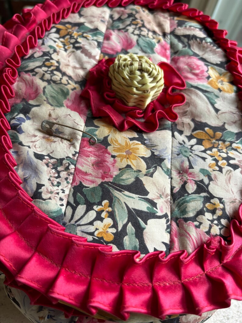 May include: A round, quilted fabric box with a floral print in shades of pink, green, and yellow. The box has a red ruffled trim and a woven straw-like knob on the top.