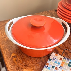 May include: A round orange casserole dish with a matching lid and white integrated side handles. The dish features a smooth, glossy finish and a central knob on the lid. Beside it sits a square trivet decorated with a multi-colored mosaic tile pattern.
