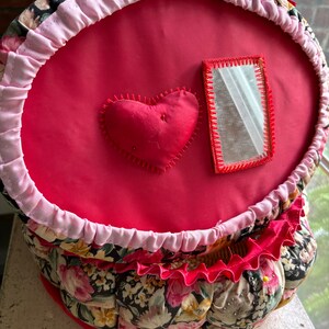 May include: A red fabric sewing basket with a pink ruffle around the top. The basket has a heart-shaped cushion and a small mirror on the front. The basket is decorated with a floral print.
