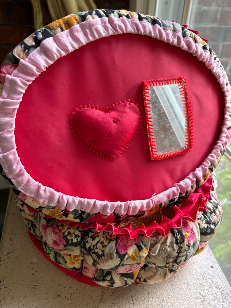May include: A red fabric sewing basket with a pink ruffle around the top. The basket has a heart-shaped cushion and a small mirror on the front. The basket is decorated with a floral print.