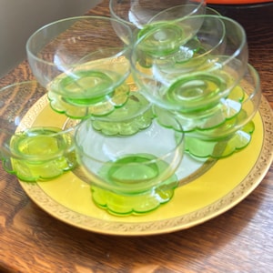May include: A collection of clear glass dessert bowls with bright green scalloped bases arranged on a round yellow plate with a gold rim. The bowls feature a rounded shape and a distinct green pedestal base, creating a layered display of glassware.