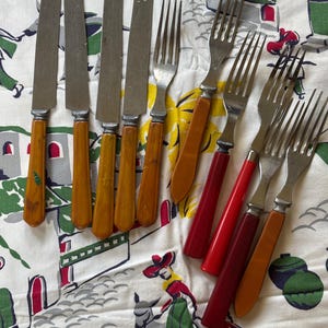 May include: A collection of vintage cutlery, including knives and forks, with handles in shades of amber, orange, and red. The stainless steel blades and tines contrast with the colorful handles. The utensils are arranged on a patterned tablecloth.