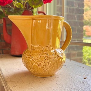 May include: A glossy mustard yellow ceramic pitcher featuring a prominent embossed relief pattern of grape clusters and vine leaves encircling the lower body. The vessel has a wide pouring spout, a curved handle, and a smooth, glazed finish.