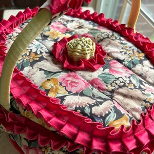 May include: A vintage floral fabric basket with a red ruffled trim and a woven straw handle. The basket has a floral pattern with pink, yellow, and green flowers on a black background.
