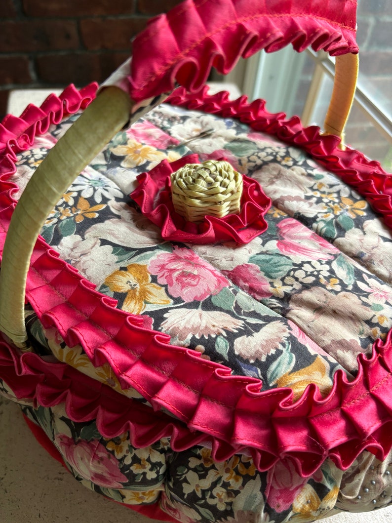 May include: A vintage floral fabric basket with a red ruffled trim and a woven straw handle. The basket has a floral pattern with pink, yellow, and green flowers on a black background.
