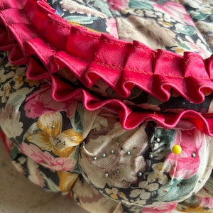 May include: A round, floral patterned cushion with a pink ruffled trim. The cushion is covered in a fabric with a variety of pink, yellow, and green flowers on a black background. There are small silver pins scattered across the surface of the cushion.
