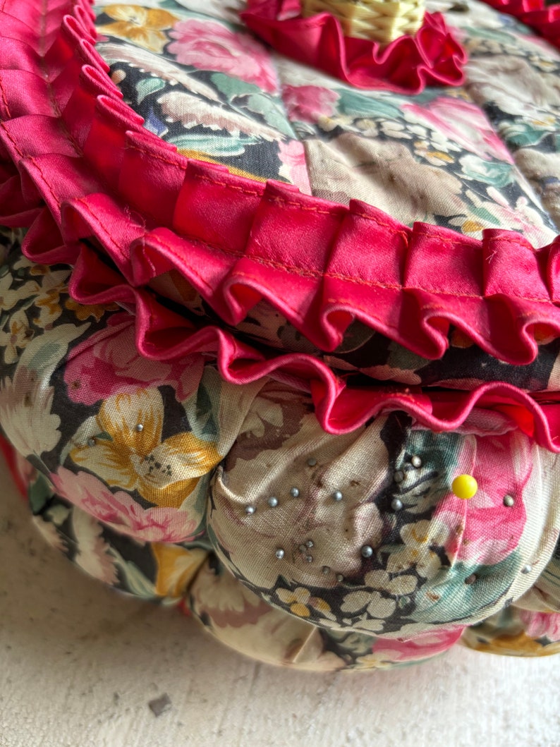 May include: A round, floral patterned cushion with a pink ruffled trim. The cushion is covered in a fabric with a variety of pink, yellow, and green flowers on a black background. There are small silver pins scattered across the surface of the cushion.