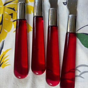 May include: Four vintage butter knives with stainless steel blades and vibrant red handles. The handles are a translucent, glossy red, and the knives are arranged on a patterned fabric background.