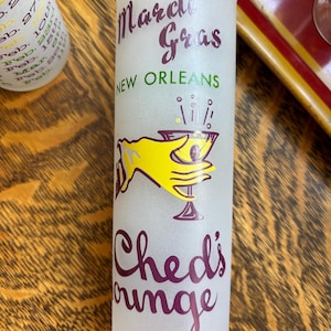 May include: Frosted glass shot glass with the words "Mardi Gras New Orleans" in purple and green. The glass features a yellow hand holding a cocktail and the words "Ched's Lounge" in purple.