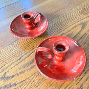 May include: Two red candle holders with a circular base and a small handle. The candle holders have a glossy finish and are sitting on a wooden surface. The candle holders are a vintage style.