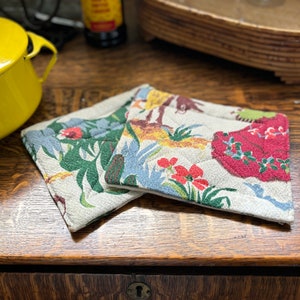 May include: Two fabric pot holders with floral and animal print designs. The pot holders are on a wooden surface.