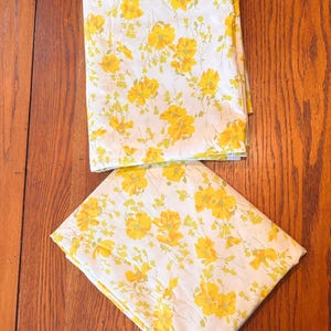 May include: Two folded pieces of white fabric featuring a repeating floral pattern in shades of bright yellow and muted green. The textile displays various sizes of blossoms and thin stems scattered across the surface in a consistent botanical print.