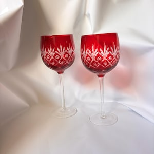May include: Two wine glasses featuring deep red bowls with clear cut glass detailing. The bowls display a diamond pattern with star-like accents etched into the surface. Each glass has a slender, clear stem and a circular base for stability.
