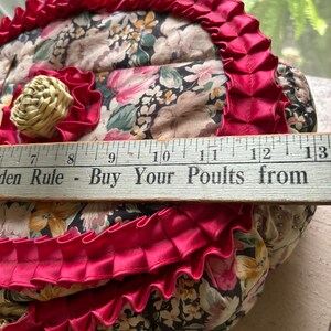 May include: A heart-shaped floral fabric basket with a pink ruffled trim and a wooden handle. The basket is decorated with a small straw flower and has a ruler with the text "Garden Rule - Buy Your Poults from" visible at the bottom.