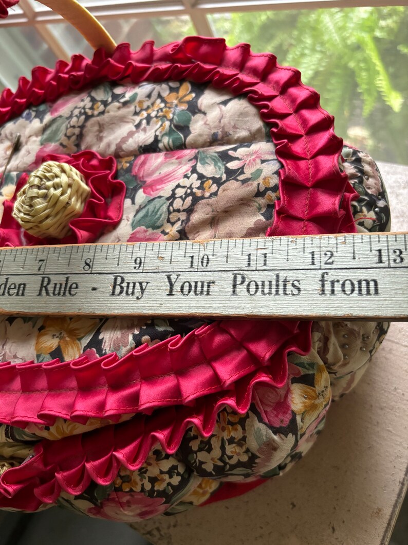 May include: A heart-shaped floral fabric basket with a pink ruffled trim and a wooden handle. The basket is decorated with a small straw flower and has a ruler with the text "Garden Rule - Buy Your Poults from" visible at the bottom.