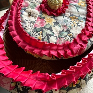 May include: A vintage wicker sewing basket with a floral fabric lining and a red ruffled trim. The lid has a small fabric flower with a straw center.
