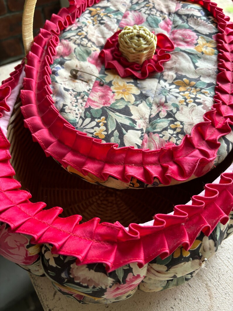 May include: A vintage wicker sewing basket with a floral fabric lining and a red ruffled trim. The lid has a small fabric flower with a straw center.