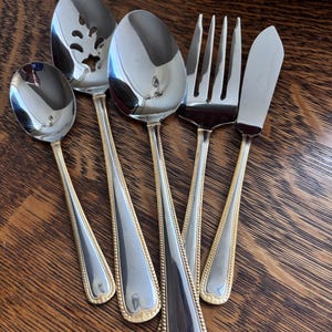 May include: A set of six silver-colored serving utensils with gold-colored trim. The set includes a spoon, a slotted spoon, a serving spoon, a fork, and a butter knife. The handles have a beaded design.