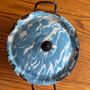 May include: A round, blue and white enamelware pot with a black handle and lid. The pot has a marbled design with a black knob on the lid. The pot is sitting on a wooden surface.