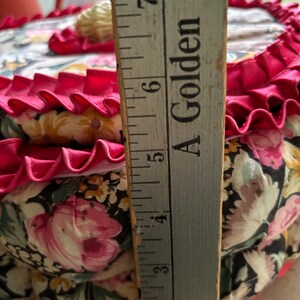 May include: A round, floral fabric pincushion with a pink ruffle trim and a white shell button on top. The pincushion is approximately 6 inches in diameter.