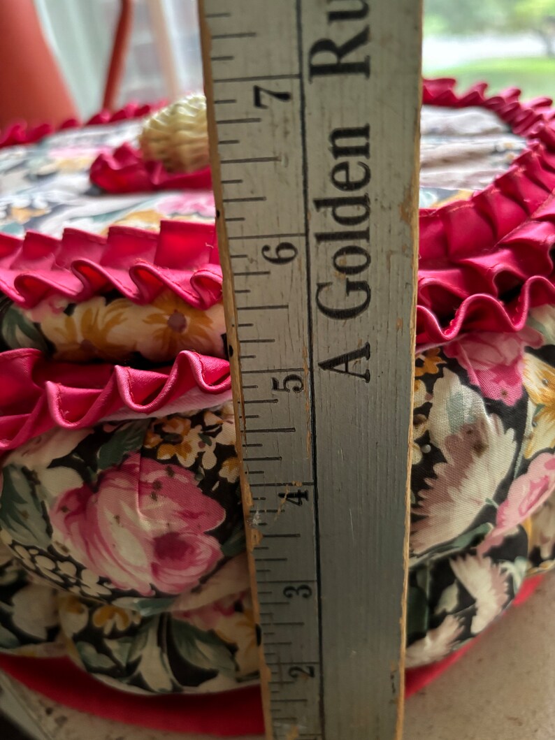 May include: A round, floral fabric pincushion with a pink ruffle trim and a white shell button on top. The pincushion is approximately 6 inches in diameter.