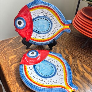 May include: Two ceramic fish-shaped plates. The plates are hand-painted with a red head, blue body, and yellow, white, and red accents. Each plate has a large eye and a series of small white lines. The plates are on a wooden table.