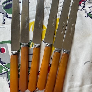 May include: A set of six stainless steel knives with orange-brown handles. The blades are silver and marked "STAINLESS STEEL". The handles have a faceted, geometric design. The knives are arranged on a patterned fabric.