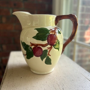 May include: Cream-colored ceramic pitcher with a brown handle and a spout. The pitcher is decorated with a painted design of red apples and green leaves on a branch. The pitcher is sitting on a white surface.