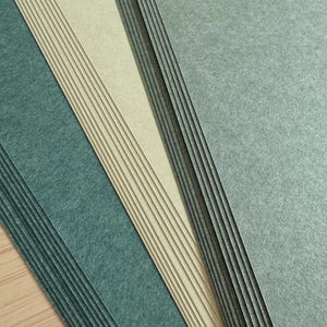 Könnte beinhalten: Ein Stapel strukturiertes Papier in Grün- und Beigetönen. Das Papier hat eine leicht raue Oberfläche und ist in einem geschichteten Muster angeordnet, wobei die Kanten jedes Blattes sichtbar sind. Die Farben reichen von einem tiefen Teal bis zu einem hellen Beige.