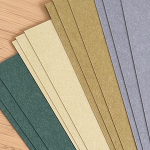 May include: A selection of rectangular paper sheets in shades of green, beige, tan, and grey. The sheets are fanned out, displaying their different colours and textures. The background is a light brown wooden surface.