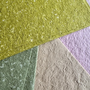Könnte beinhalten: Eine Sammlung handgefertigter Papierbögen in verschiedenen Farben. Die Bögen haben ein strukturiertes, faseriges Aussehen mit weißen Flecken. Zu den Farben gehören Grün-, Lavendel- und Beigetöne. Das Papier scheint dick und für Bastelarbeiten geeignet zu sein.